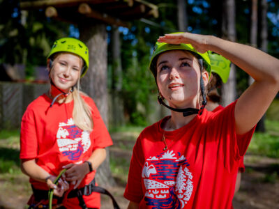 Dwie dziewczyny w kaskach i czerwonych koszulkach na tle parku linowego.