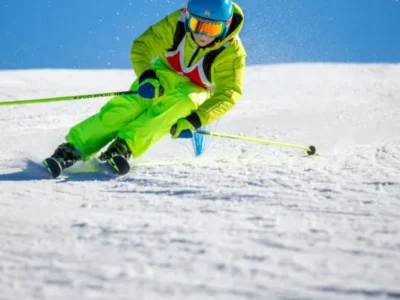 Młody narciarz w jaskrawej kurtce zjeżdża po stoku, tworząc śnieżny obłok.