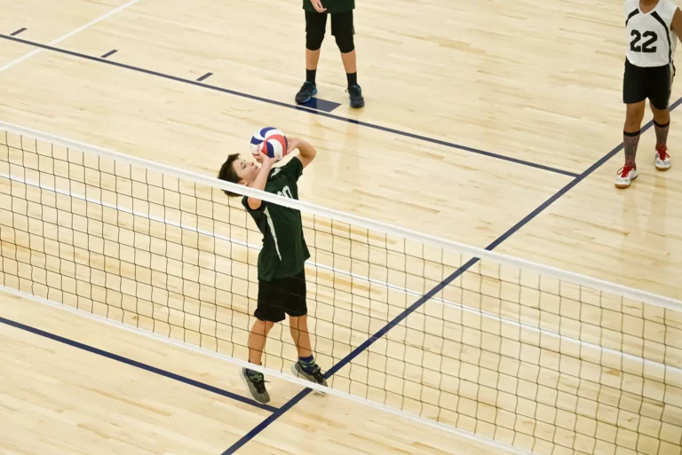 Chłopiec w zielonej koszulce serwuje piłkę do siatkówki na drewnianej hali sportowej.