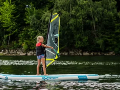 Dziewczynka na desce windsurfingowej, nauka żeglowania, aktywność wodna, wakacje na jeziorze.