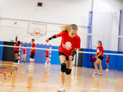 Dziewczynka w czerwonej koszulce ćwiczy siatkówkę na hali sportowej.