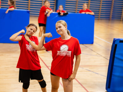 Dwie dziewczynki w czerwonych koszulkach z logo siatkówki pokazują siłę na sali gimnastycznej.