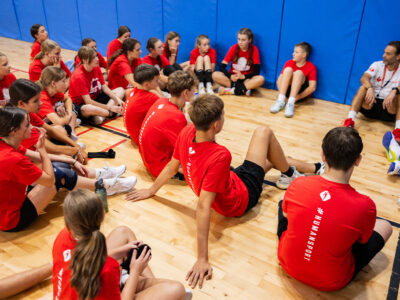 Dzieci w czerwonych koszulkach (#HUMANSPORT) siedzą na podłodze, słuchając trenera.
