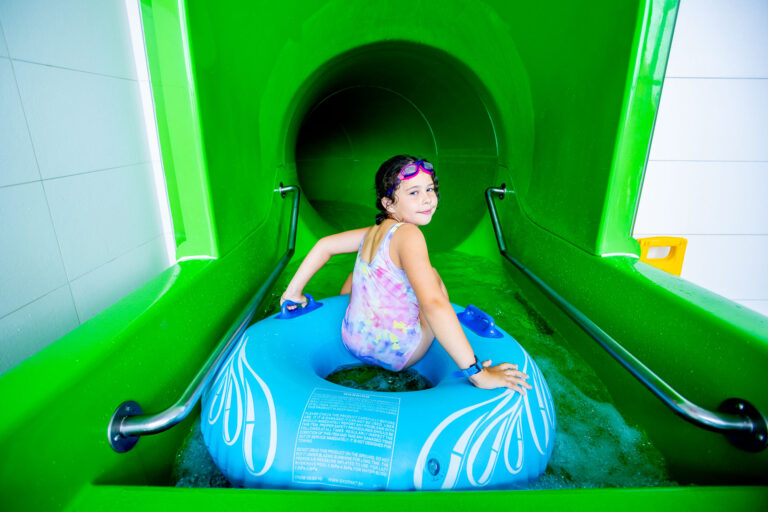 Dziewczynka z kółkiem zjeżdża po zielonej zjeżdżalni wodnej.