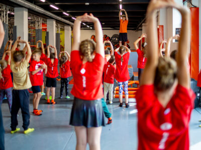 Dzieci w czerwonych koszulkach ćwiczą z trenerem w sali gimnastycznej.
