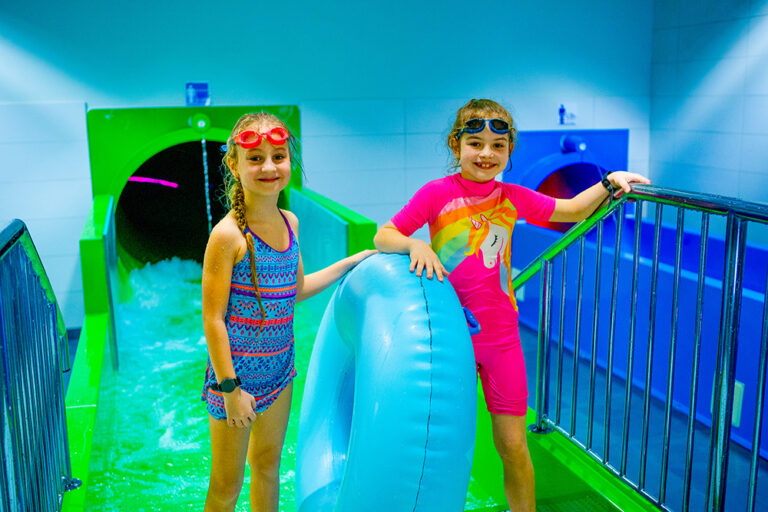Dwie dziewczynki w strojach kąpielowych i goglach na głowach z niebieską dmuchaną zabawką w aquaparku.