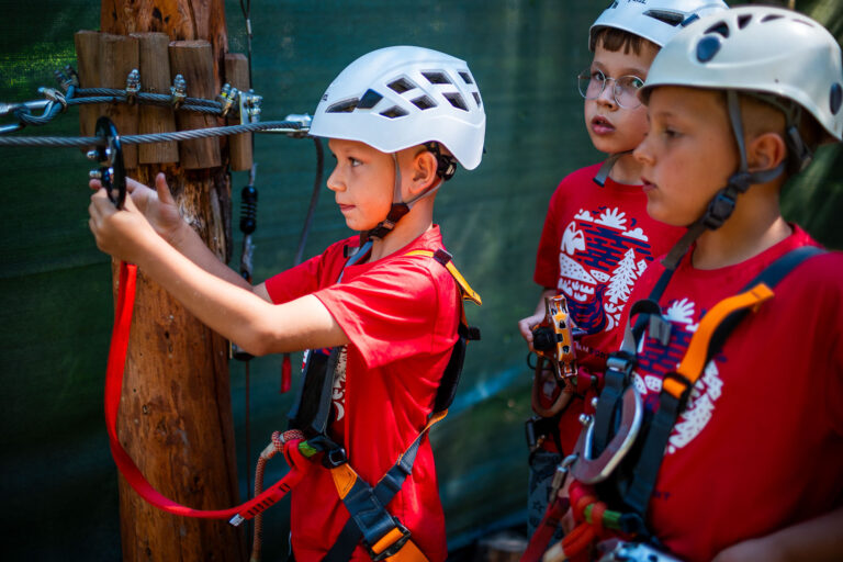 Dzieci w kaskach i szelkach na ściężce linowej. Przygoda i zabawa na wysokości.