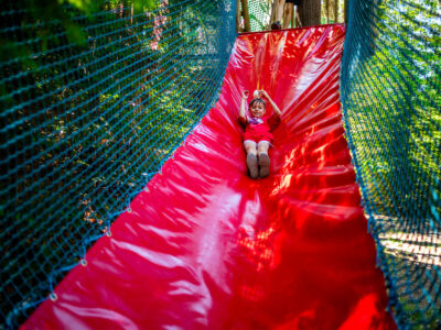 Dziecko zjeżdża po czerwonej zjeżdżalni w parku linowym.