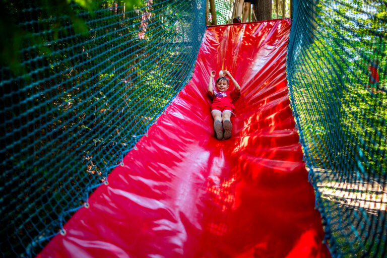 Dziecko zjeżdża po czerwonej zjeżdżalni w parku linowym.
