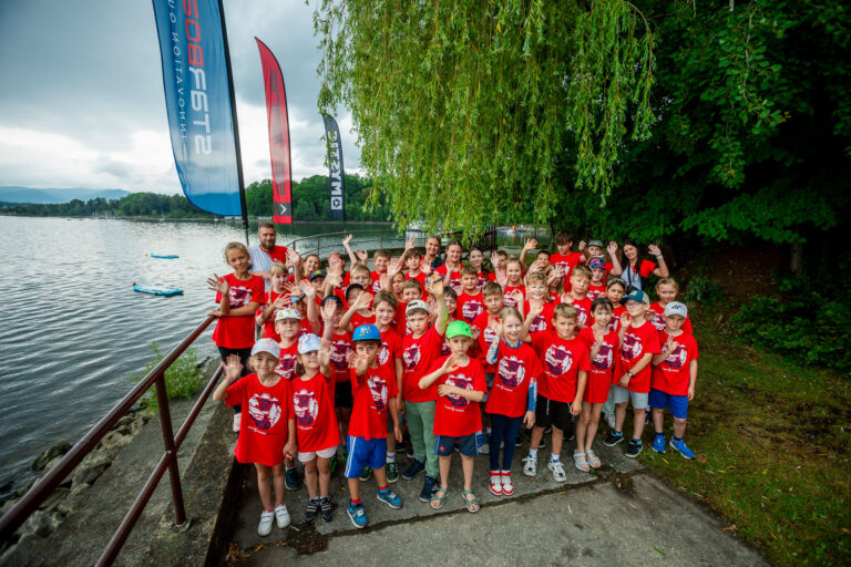 Grupa dzieci w czerwonych koszulkach na tle jeziora i drzew.