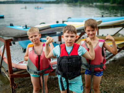 Trzech chłopców w kamizelkach ratunkowych pozuje z uśmiechem nad jeziorem, gotowi na wodne przygody.