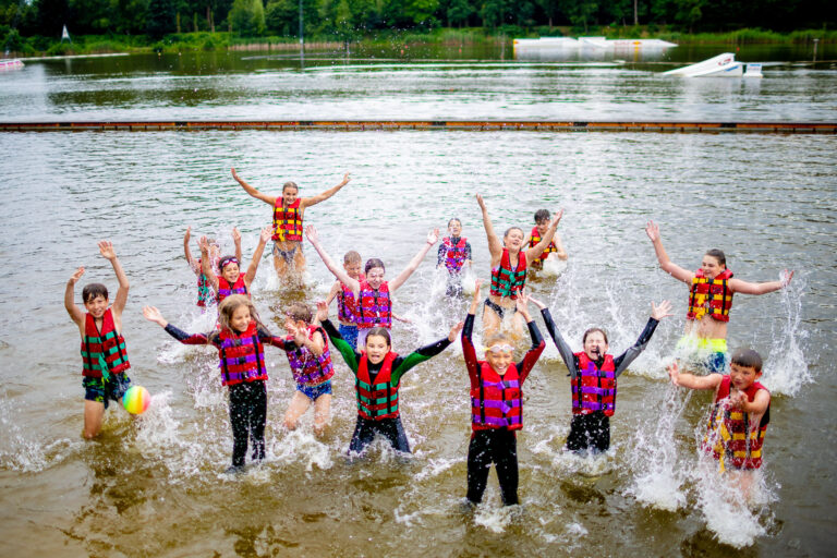 Dzieci w kamizelkach ratunkowych pluskają się w wodzie, radośnie unosząc ręce.