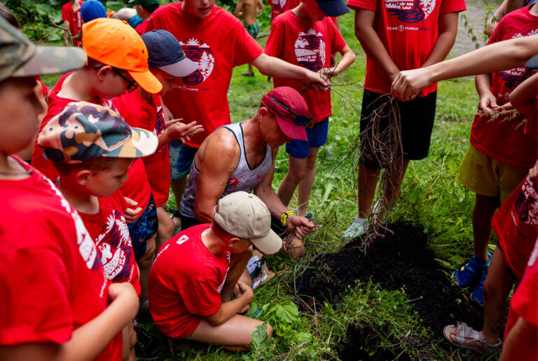 Dzieci w czerwonych koszulkach i czapkach obserwują mężczyznę pracującego z ziemią i gałązkami.
