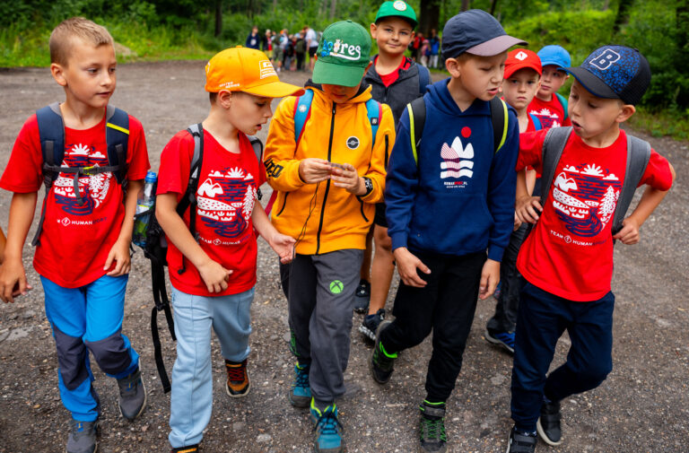 Grupa dzieci w kolorowych ubraniach i czapkach idzie leśną ścieżką.