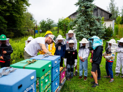 Dzieci w strojach pszczelarskich obserwują pracę pszczelarza przy ulu. Edukacja o pszczołach.