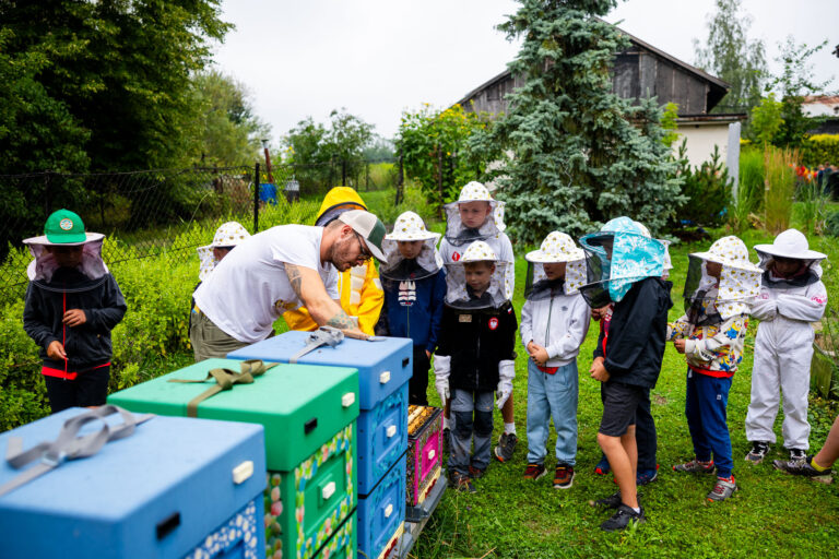 Dzieci w strojach pszczelarskich obserwują pracę pszczelarza przy ulu. Edukacja o pszczołach.