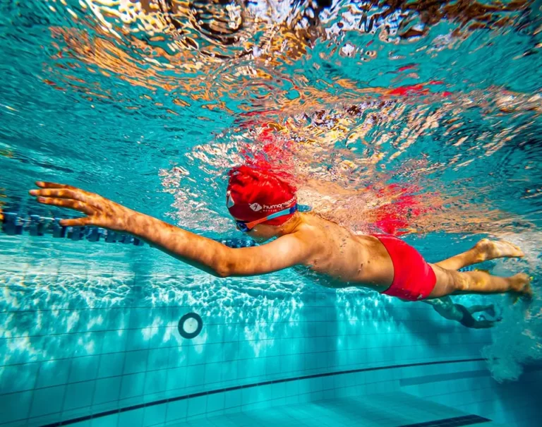 Chłopiec w czerwonym czepku pływa pod wodą.