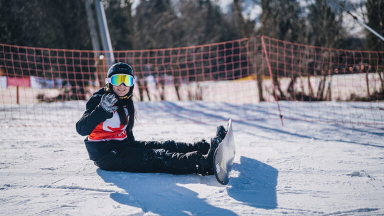 Snowboardzistka w okularach siedzi na śniegu, machając ręką.