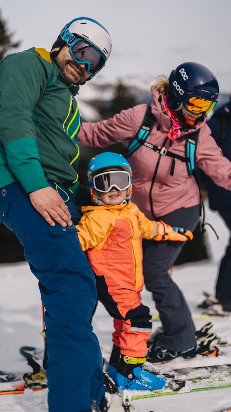 Rodzina na nartach: uśmiechnięte dziecko w pomarańczowej kurtce i rodzice w tle.