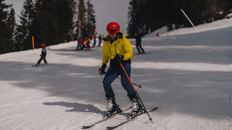 Narciarz w żółtej kurtce zjeżdża po stoku. Narty, kije, kask, góry.