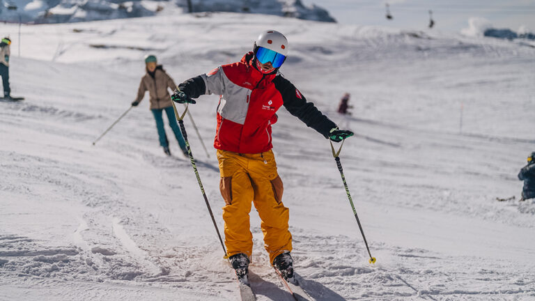 Narciarz w jaskrawym stroju zjeżdża po ośnieżonym stoku, trzymając kije.