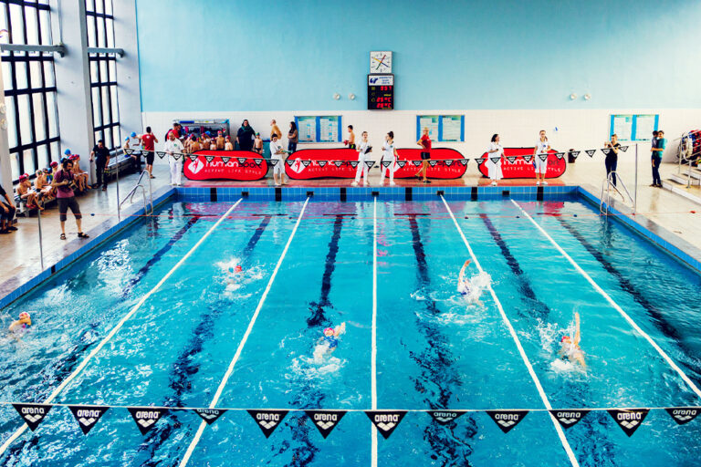 Zawody pływackie na basenie, dzieci i dorośli rywalizują w wyścigach.